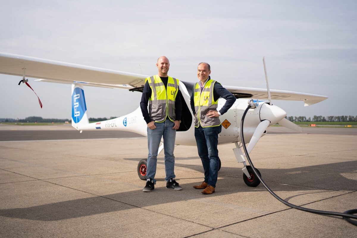 Twee mannen in gele hesjes poseren voor een klein elektrisch vliegtuig dat met een snoer wordt opgeladen.